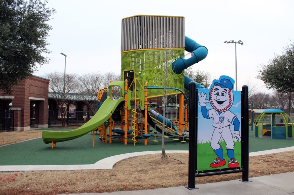A playground behind a sign featuring imagery of a cartoon mascot with a baseball head wearing a white and blue Miracle League baseball jersey.