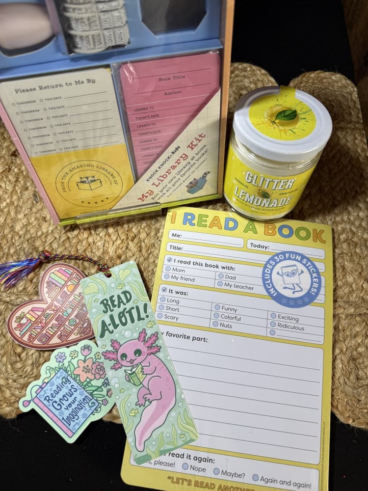 Assortment of book-themed merchandise including library checkout cards, reading log sheet, bookmarks with decorative designs, and a novelty candle arranged on woven basket surface.