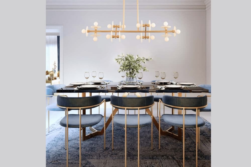 Modern linear gold chandelier hanging over an elegant dining table with blue velvet chairs.
