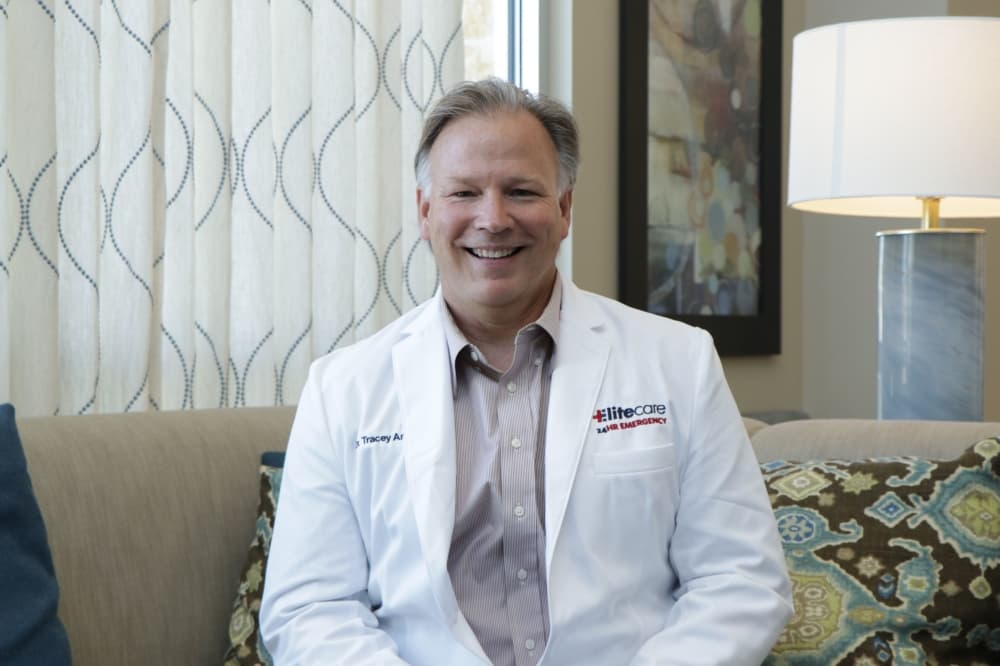 Portrait of Dr. Tracey Antill, a smiling physician in a white lab coat with the EliteCare 24HR Emergency logo, sitting in a comfortable medical office setting.