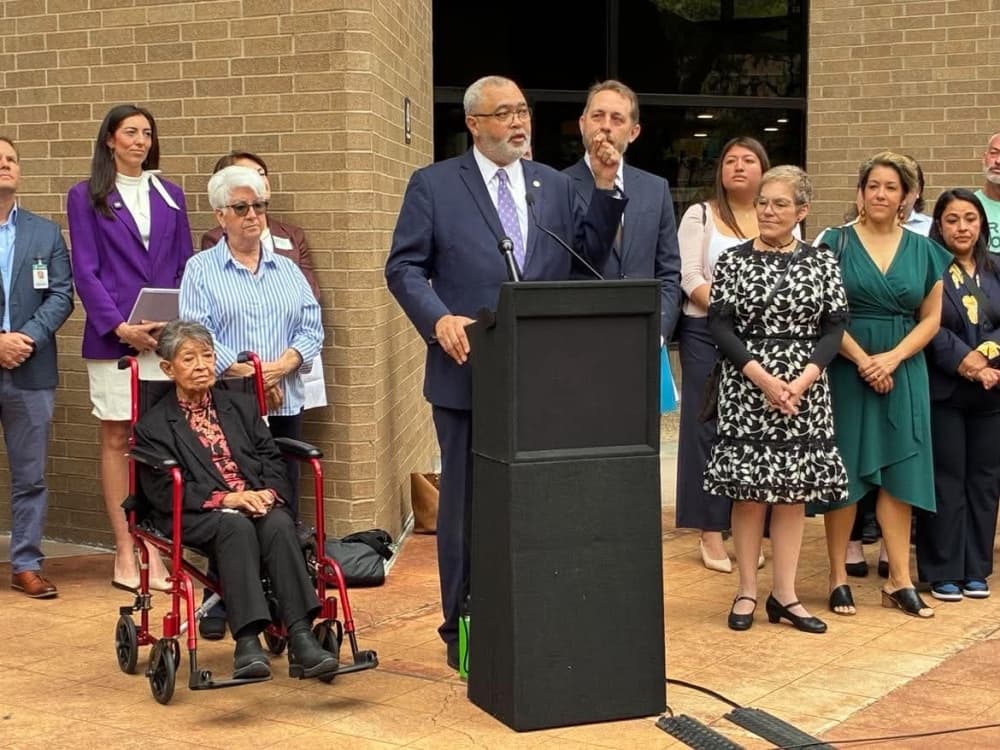 Travis County Commissioner Jeff Travillion speaks at an outdoor podium surrounded by community leaders and officials outside the Travis County Administration Building.