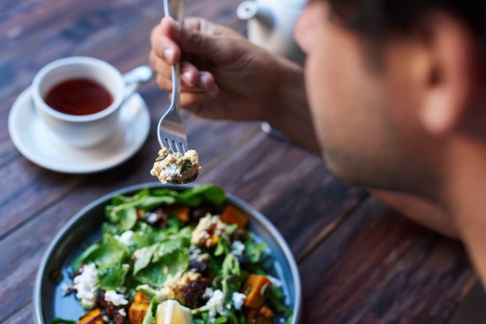 Young man sitting alone at a bistro table eating a delicious plate of mixed salad with a fork and drinking a cup of tea