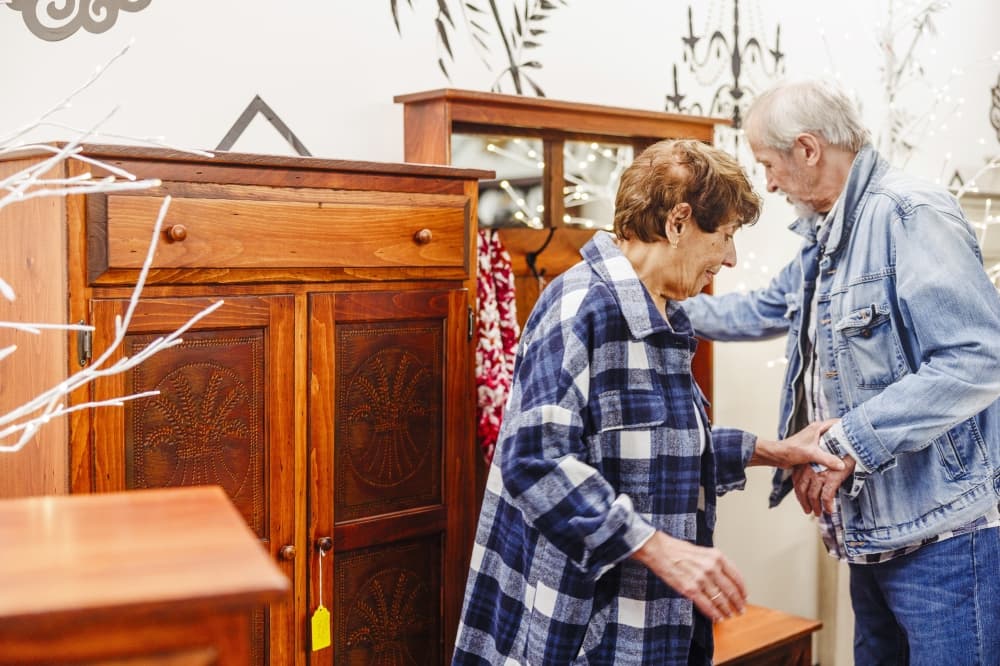 Senior couple examines wooden furniture in antique store. Woman in blue plaid shirt leans forward to inspect cabinet drawer, while male companion in denim jacket stands close by. Various vintage pieces, mirror, and decorative items surround them, creating cozy, nostalgic atmosphere typical of Poconos region shops.