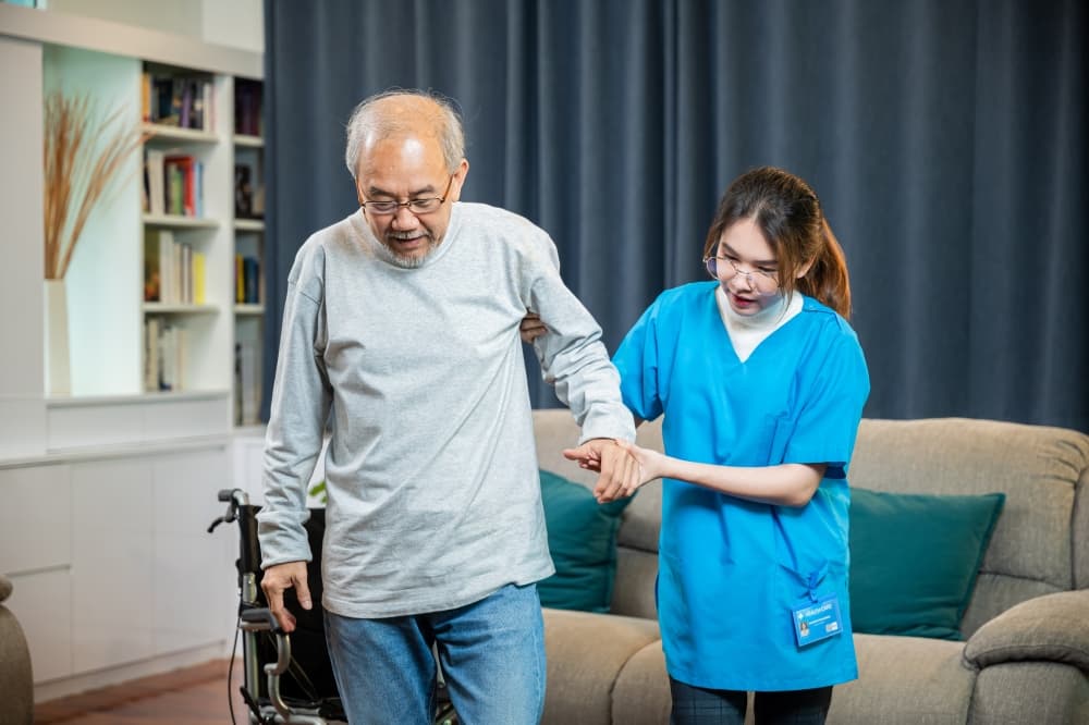 doctor support old man to getting up to exercise, help handicapped elderly stand up, woman nurse assisting helping senior man patient get up from wheelchair for practice walking at home