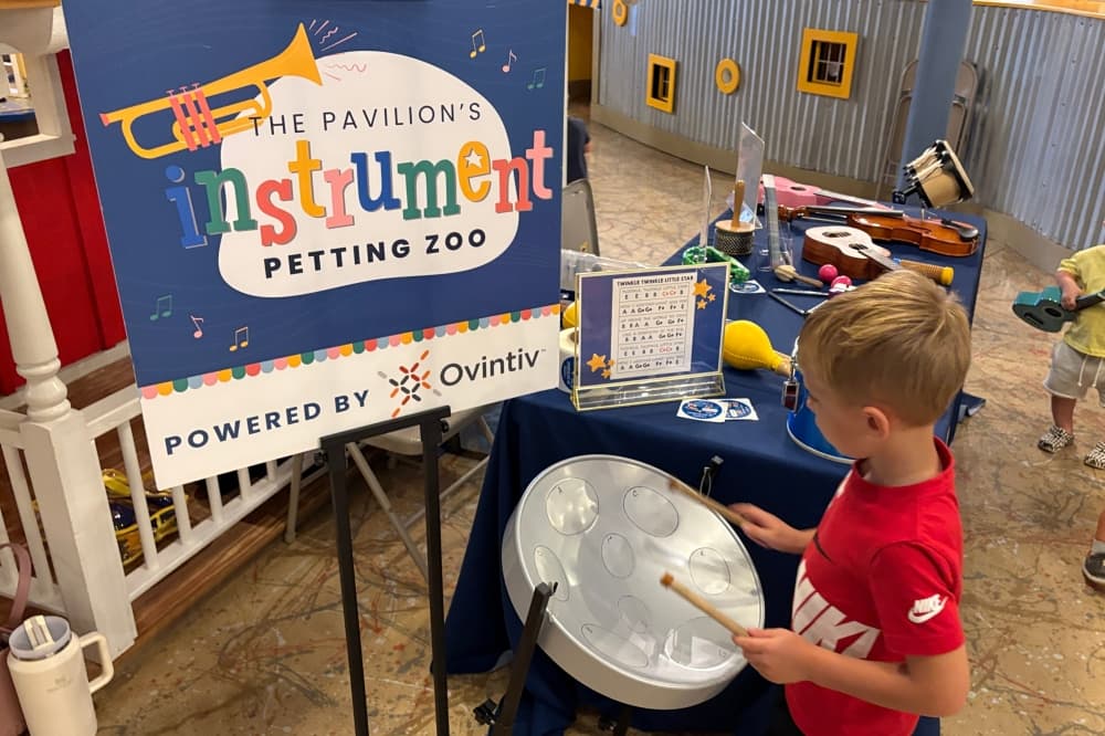 Child in a red shirt playing a drum, he's standing in front of a sign that reads 'Pavilion's Instrument Petting Zoo.'