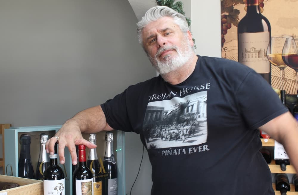 A man with a white beard in a black shirt poses with his arm resting on a bottle of wine
