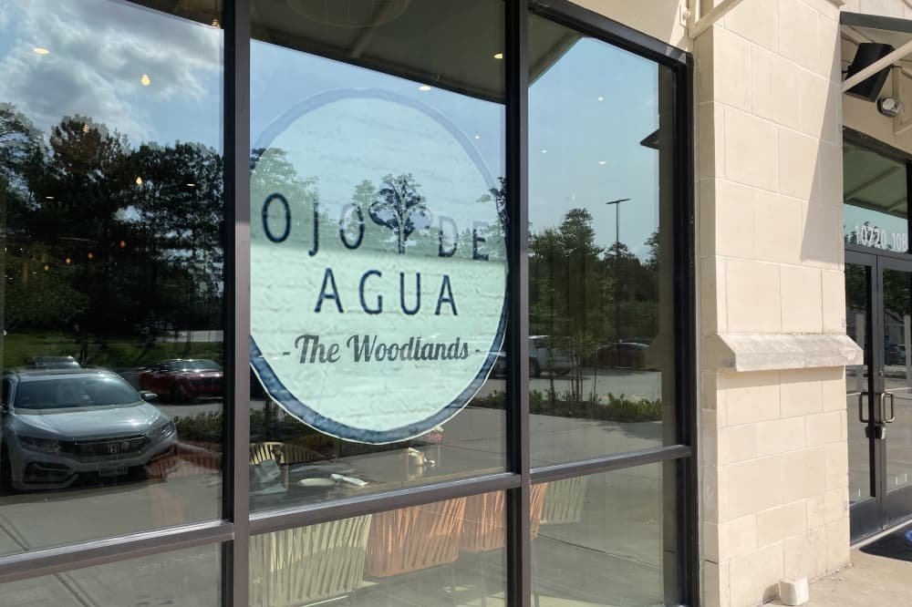 Exterior view of Ojo de Agua's window sign. The name of the eatery is written in black text and is inside a white circle.