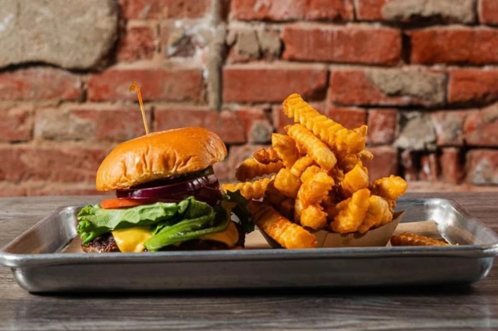 A cheeseburger with a toothpick sticking out on a metal tray next to a stack of french fries