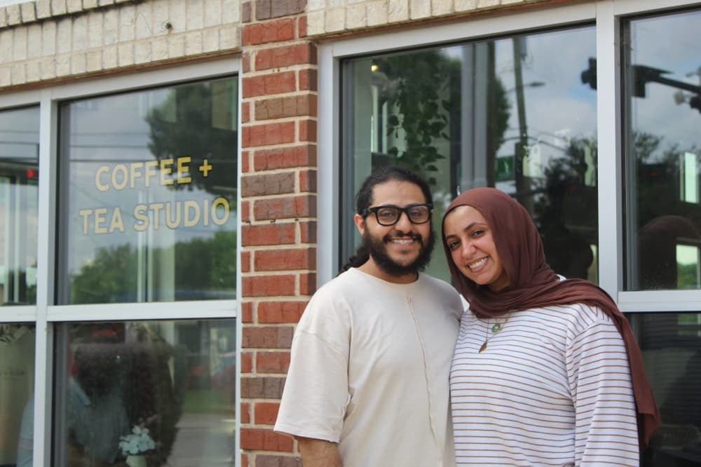 A man with glasses and a beard and a woman wearing a brown hijab stand in front of window with a decal reading "COFFEE AND TEA STUDIO"