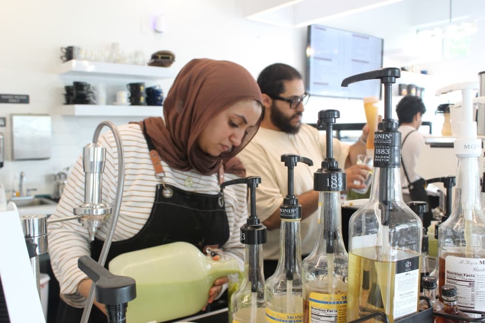 A woman in an apron pours green milk into a cup behind a row of syrup pumps