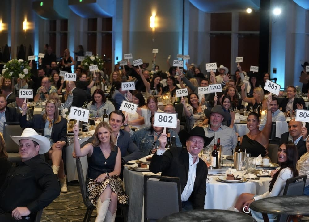 Gala attendees seated at round tables scattered throughout a venue space, each attendee is smiling at the camera and holding up auction signs with numbers on them