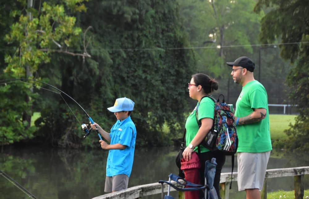 Two parents in matching green shirts spectating their child as he fishes from a lakeside