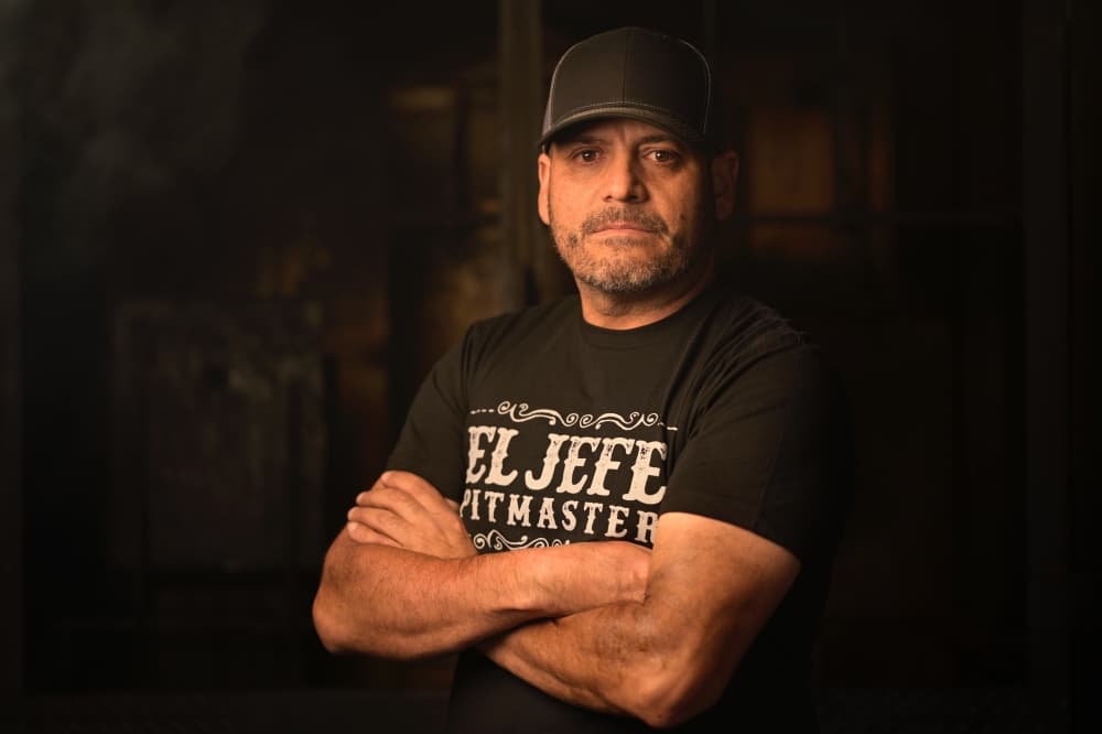 A man in a black shirt and baseball cap stands with his arms crossed
