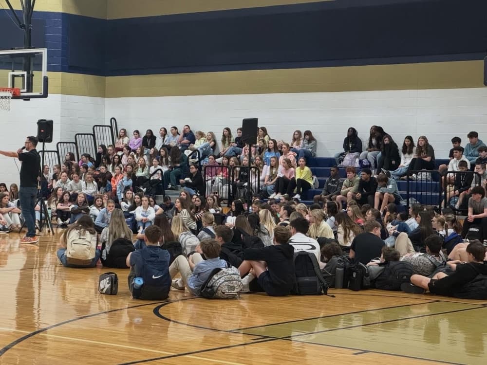 Side view of huddle event hosted in a high school gymnasium, there is a crowd of students sitting on the floor and on the bleachers