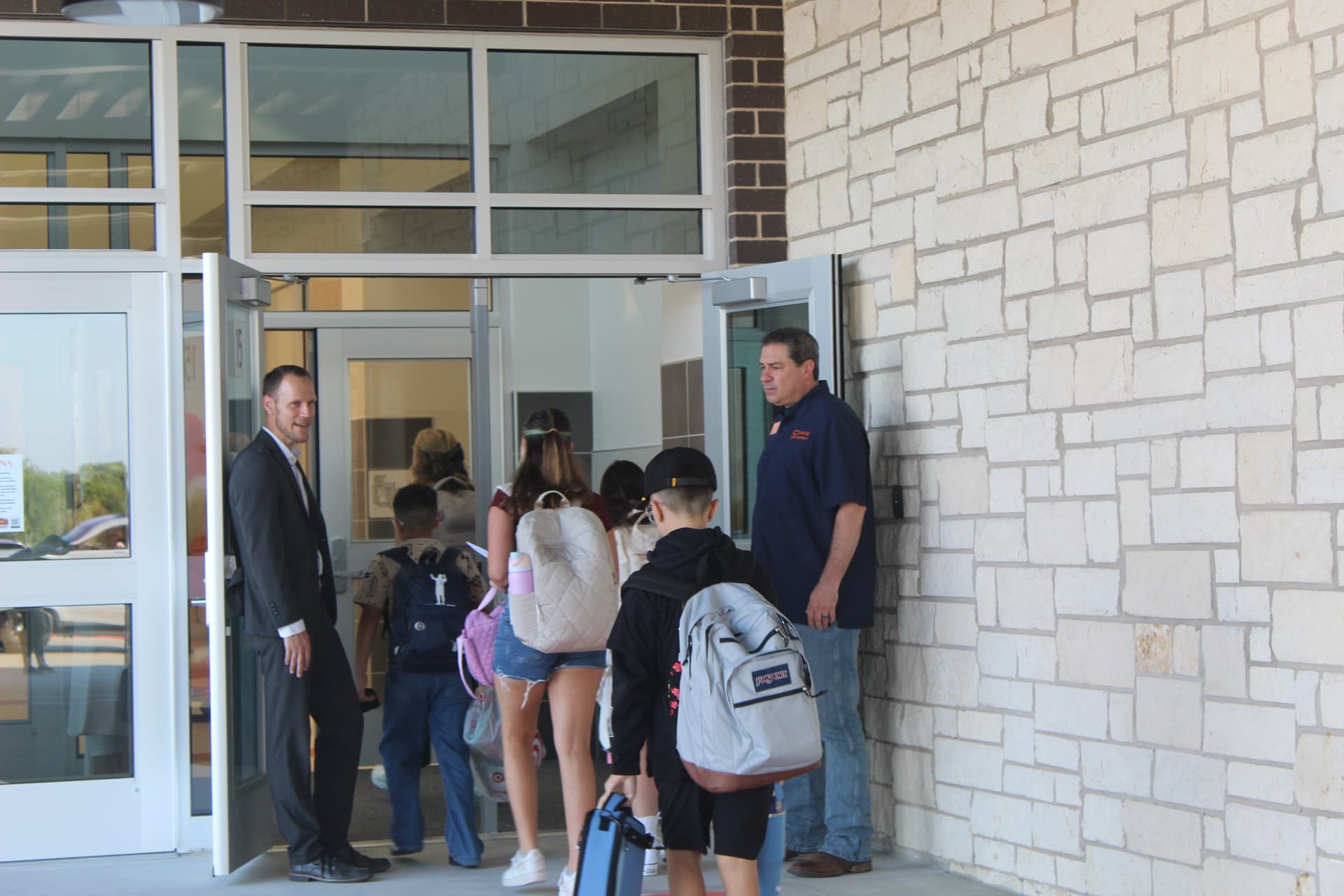 Two men guide children with backpacks through a set of glass doors