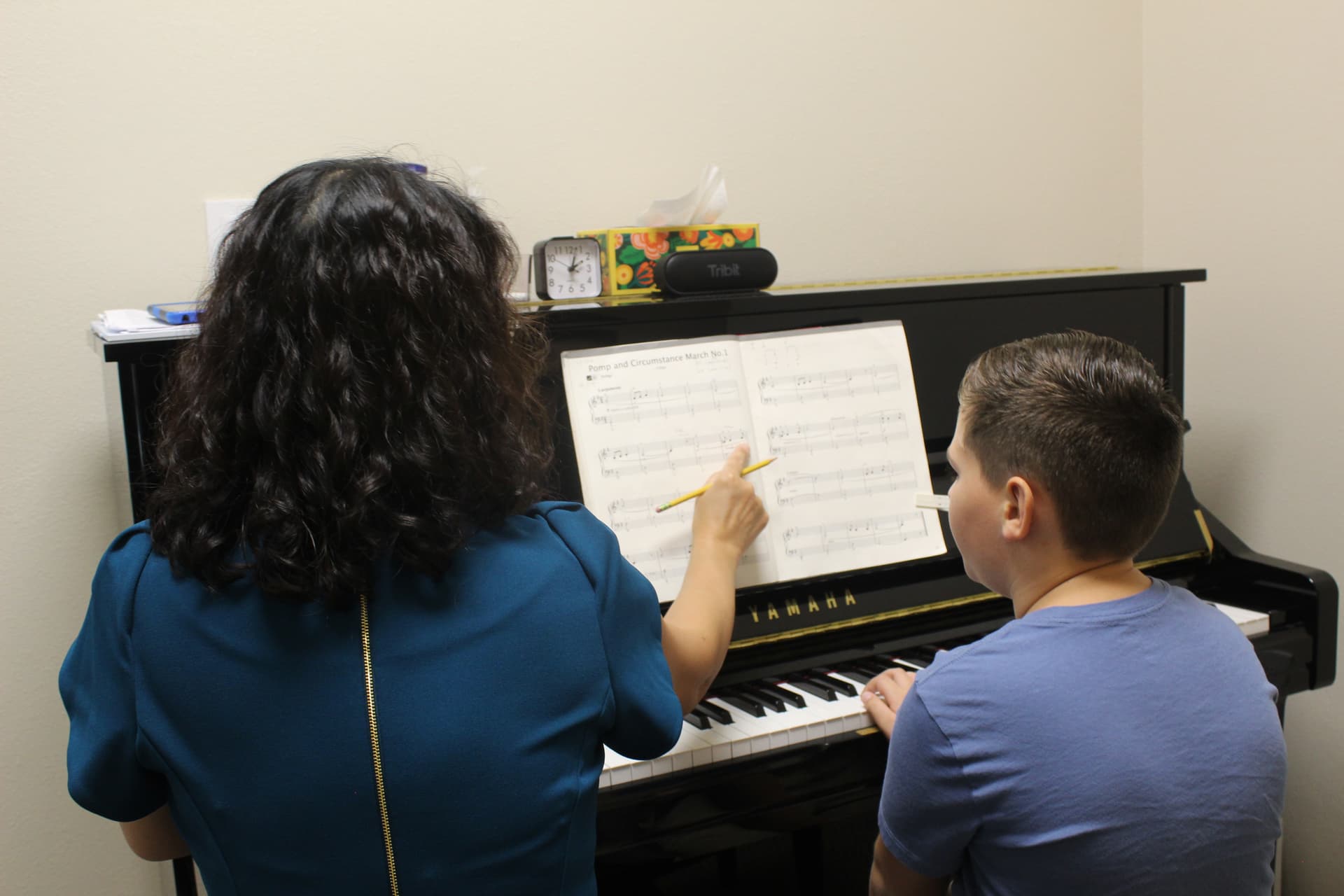 A woman in a blue dress points to sheet music for a boy in a blue shirt