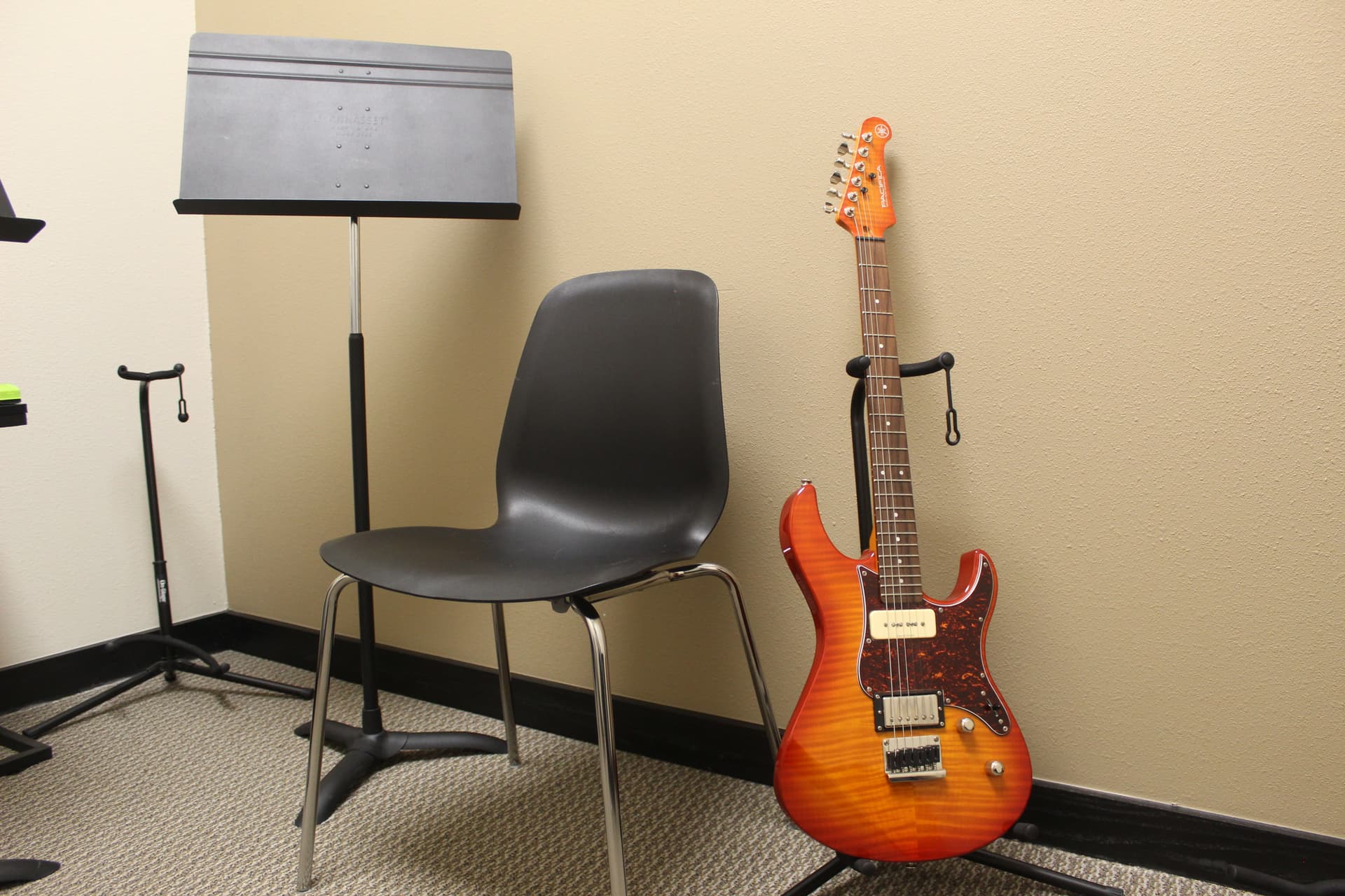 An orange electric guitar sits on a stand next to a chair and music stand