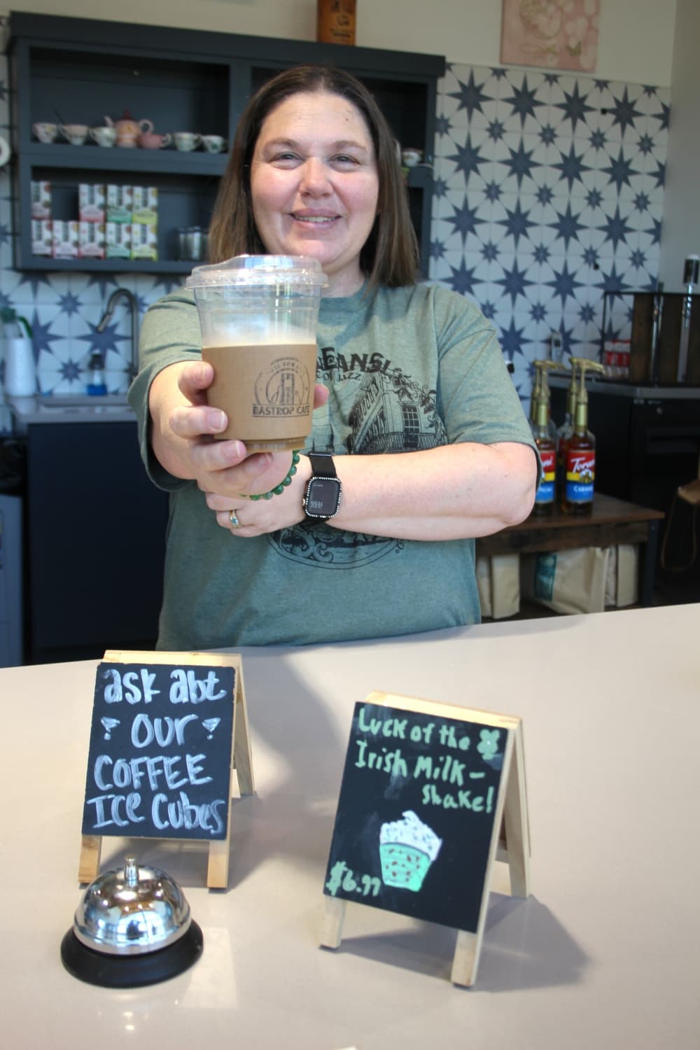 Owner Christina Want stands behind the counter smiling, while holding up a cup of iced coffee toward the camera.