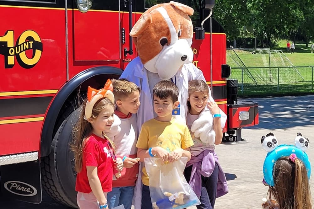 Children smile and gather around a dog mascot for the fire department in front of a fire truck.