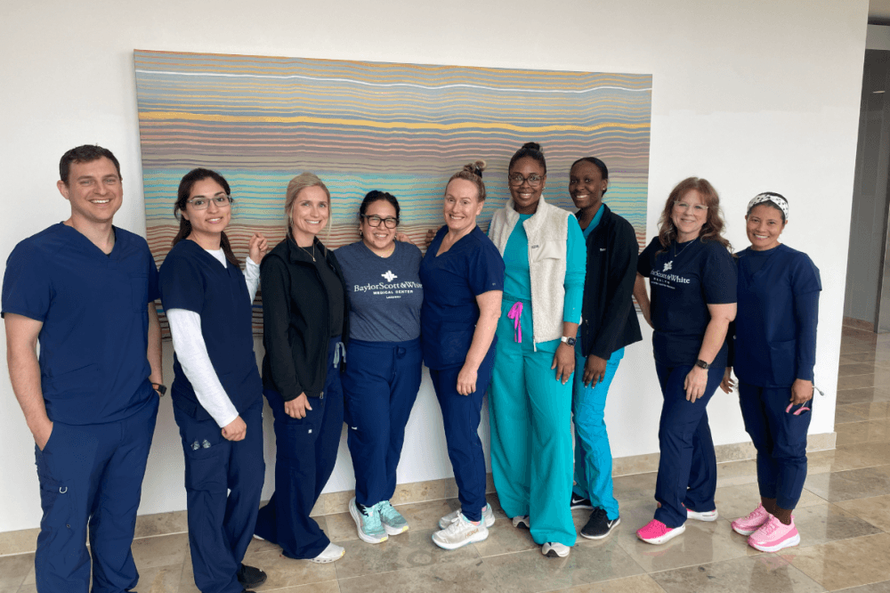 A group of nine nurses and healthcare workers in scrubs smile together inside Baylor Scott & White Medical Center Lakeway, standing in front of a colorful abstract painting.