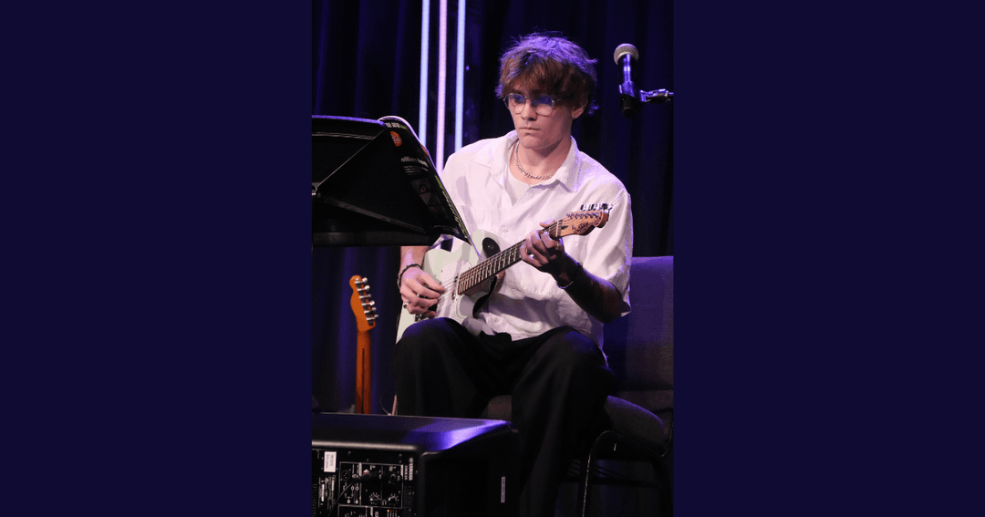 Young man in white shirt plays electric guitar seated on stage beside a music stand and microphone.