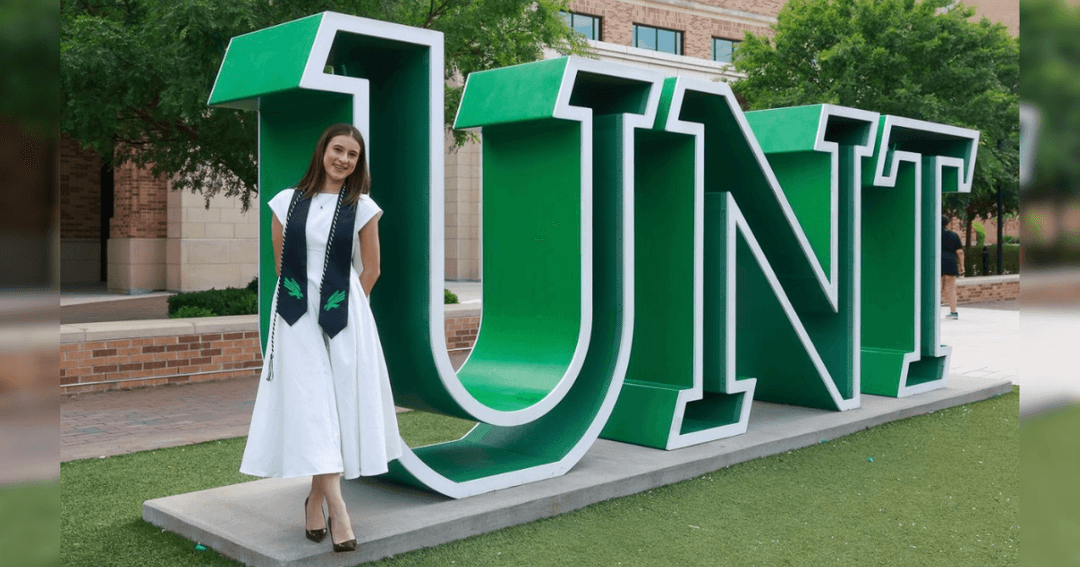Graduate in white dress with green stole poses beside large green UNT letter sculpture on campus.