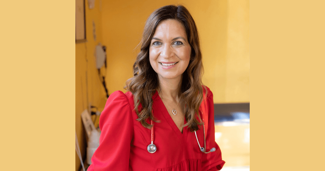 Smiling woman in red top with stethoscope around her neck, in a medical setting with yellow walls.