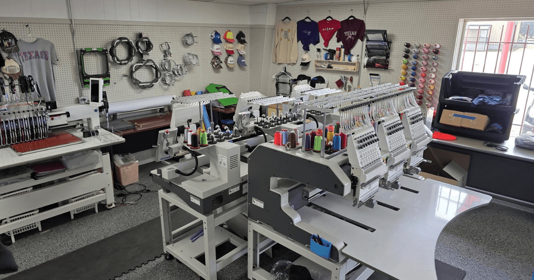 Embroidery shop with multi-head machines, colorful thread spools, and custom shirts and caps on display walls.