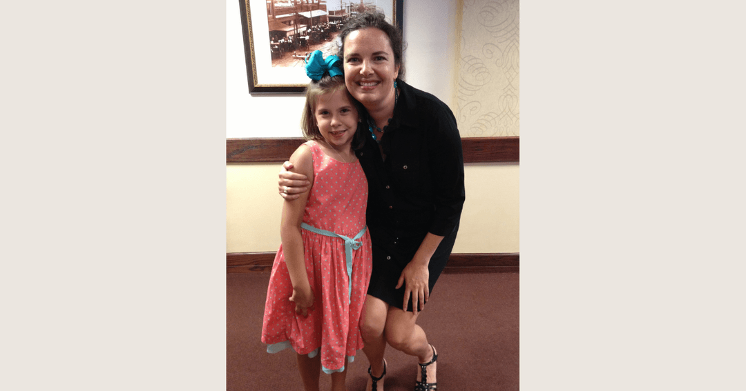 Woman in black dress kneels beside smiling young girl in pink dress with teal bow, both posing indoors.