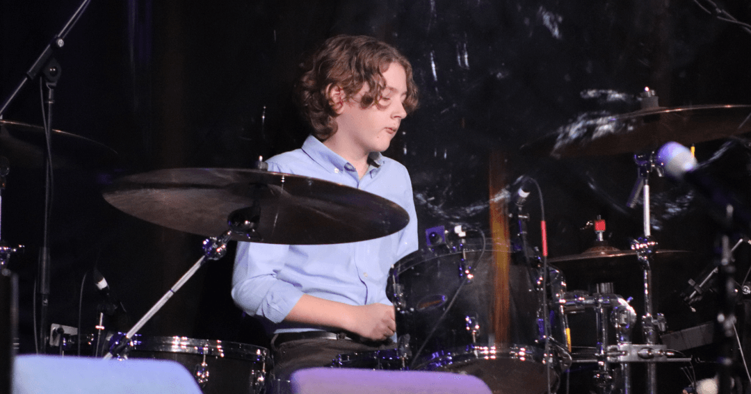 Young drummer in a blue shirt plays a drum kit on a dimly lit stage.
