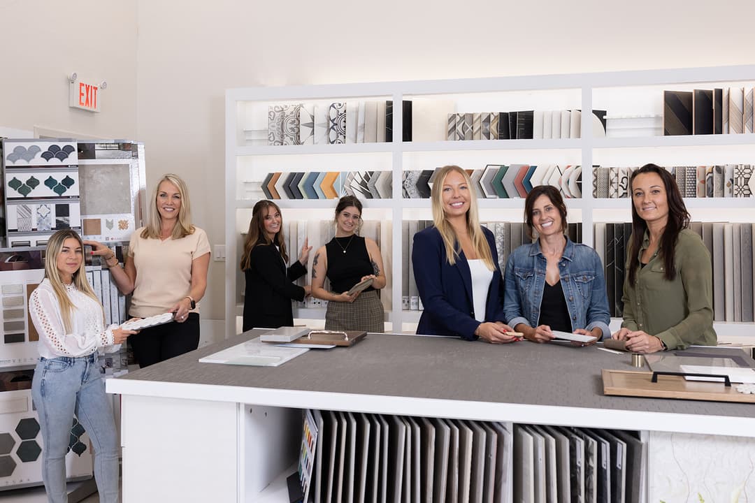 Seven women pose in a tile and design showroom with sample displays lining the walls.