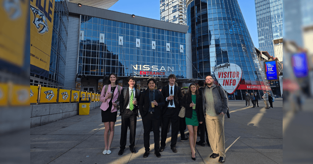 Six people in business attire pose outside Nissan Stadium entrance in Nashville near a Visitor Info sign.