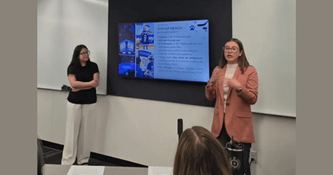 Two students give a presentation beside a screen displaying a "Pop-Up Merch" slide in a classroom.