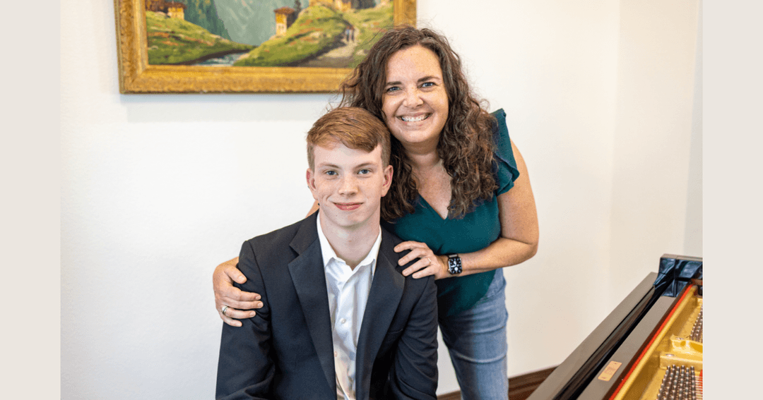 Woman and young man in suit smiling beside a grand piano, framed landscape painting on wall behind them.