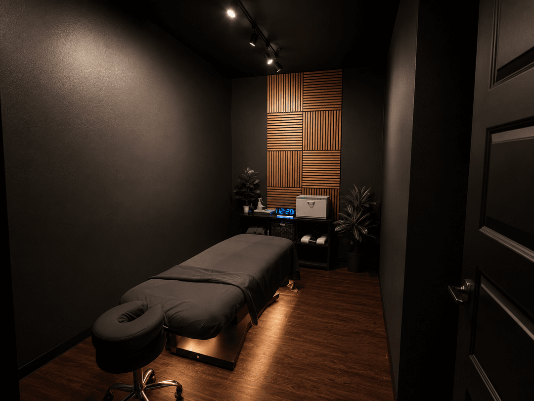 Dimly lit massage room with a treatment table, dark walls, wood accent panel, plants, and a digital clock.