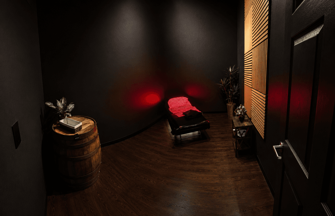 Dark spa room with red light therapy bed, wooden barrel table, plants, and slatted wood wall panel.