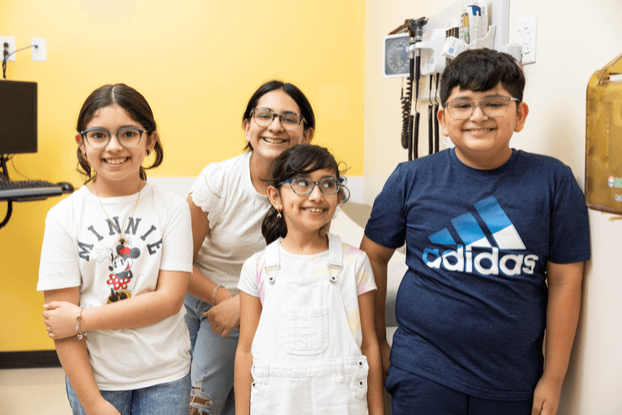 Four smiling children wearing glasses stand together in a medical exam room with yellow walls.