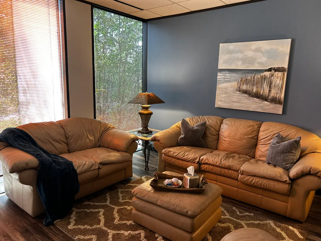 Cozy office lounge with tan leather sofas, ottoman, beach painting, lamp, and windows overlooking trees.