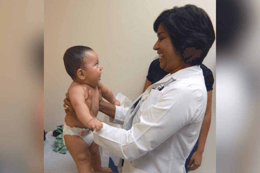 Smiling doctor in white coat holds a baby during an exam, both looking at each other.