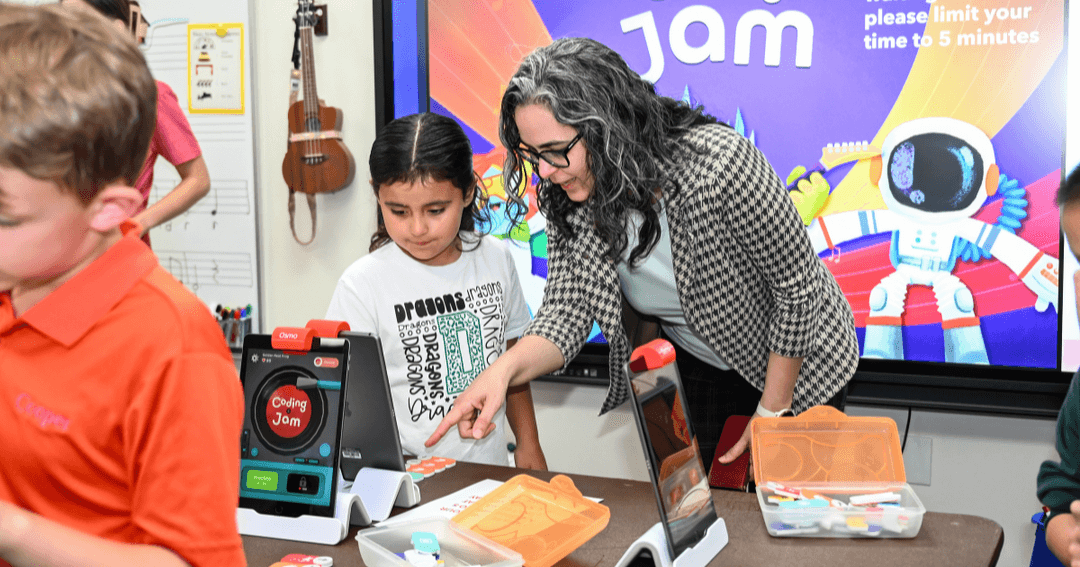 A teacher points at a tablet screen to help a student with a Coding Jam activity in a classroom.