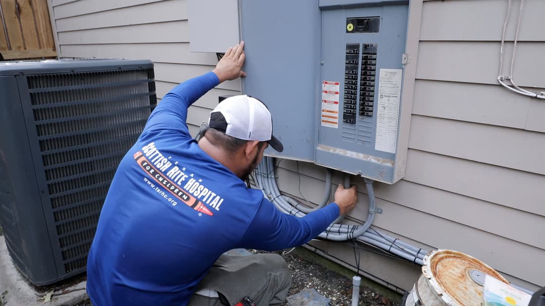 Worker in blue shirt and cap examines an open outdoor electrical breaker panel beside an AC unit.