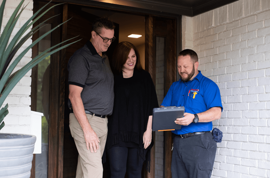 HVAC technician in blue uniform shows a tablet to a smiling couple at their front door.
