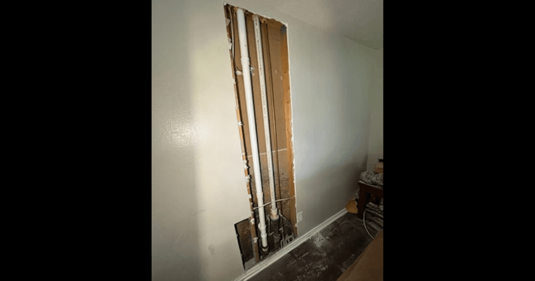 Wall opened to expose vertical plumbing pipes, with drywall removed and interior studs visible.