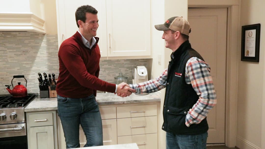 Two men shake hands and smile in a kitchen with white cabinets and a red kettle on the stove.