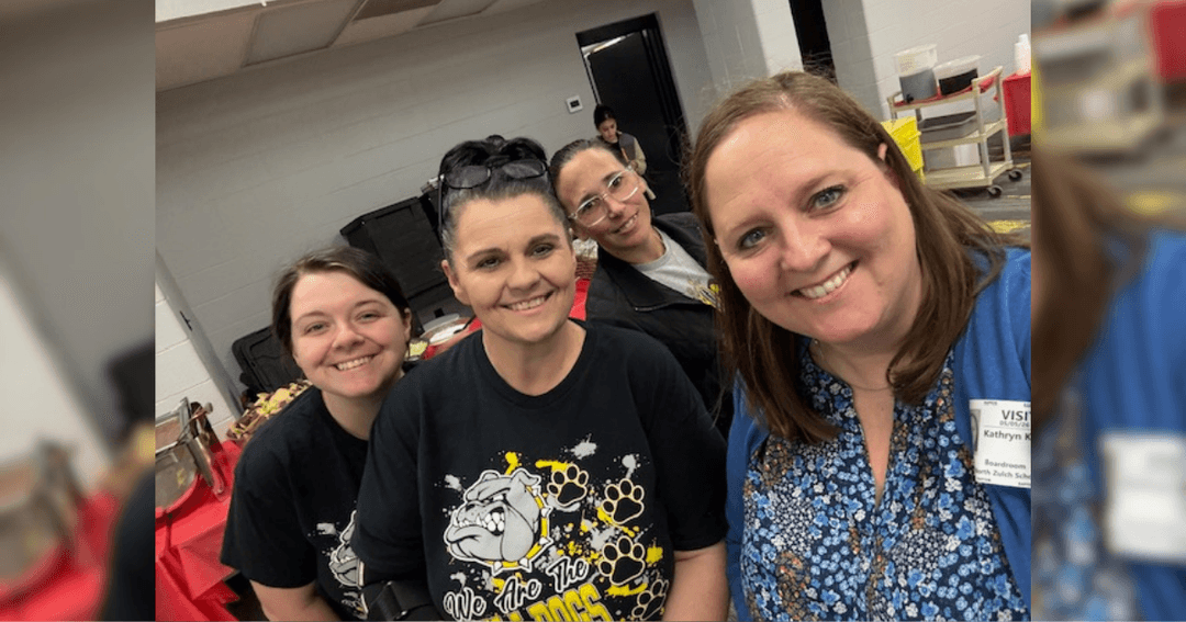Four women smiling together indoors, one wearing a Bulldogs T-shirt, another with a visitor name badge.