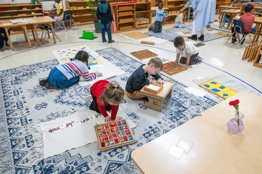 Young children work with Montessori learning materials on rugs in a classroom while a teacher assists.