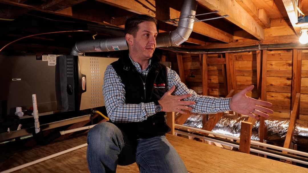 Man in vest gestures while explaining beside a Lennox furnace and ductwork in an unfinished attic or crawlspace.