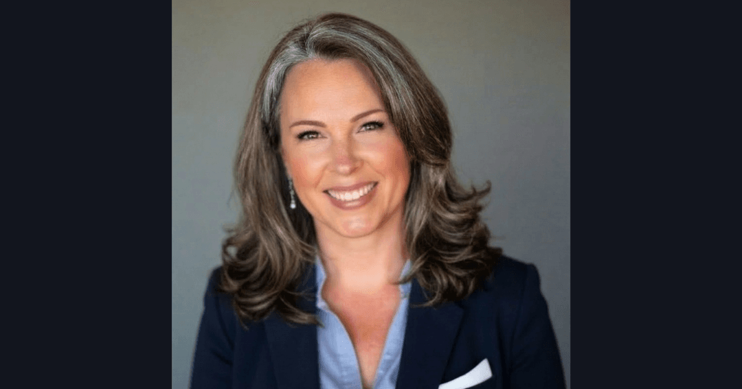 Smiling woman with wavy gray-brown hair wearing a navy blazer over a light blue shirt in a professional portrait.