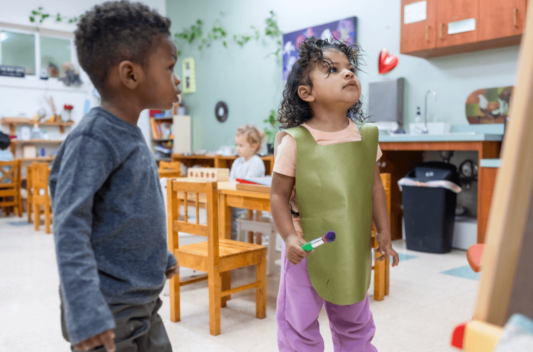 Young child in green smock holds a paintbrush in a classroom as another child looks on.