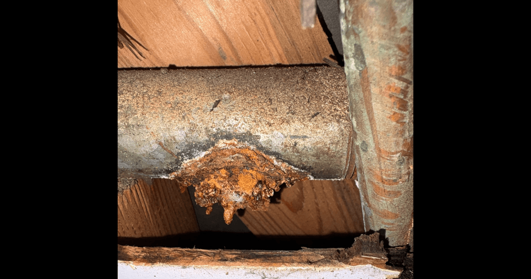 Corroded metal pipe with rust buildup and mineral deposits leaking below wooden framing.